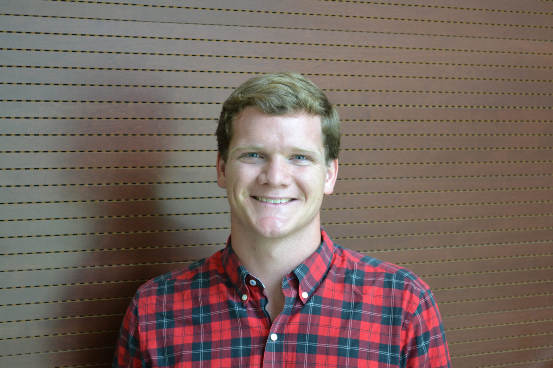 Picture of Nicholas L. Johnson. A late twenty-year old masculine presenting person with short light brown hair is wearing a red and black tartan button down shirt.
